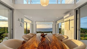 A modern dining area with a long wooden table, white chairs, and large windows offering a view of the sea.