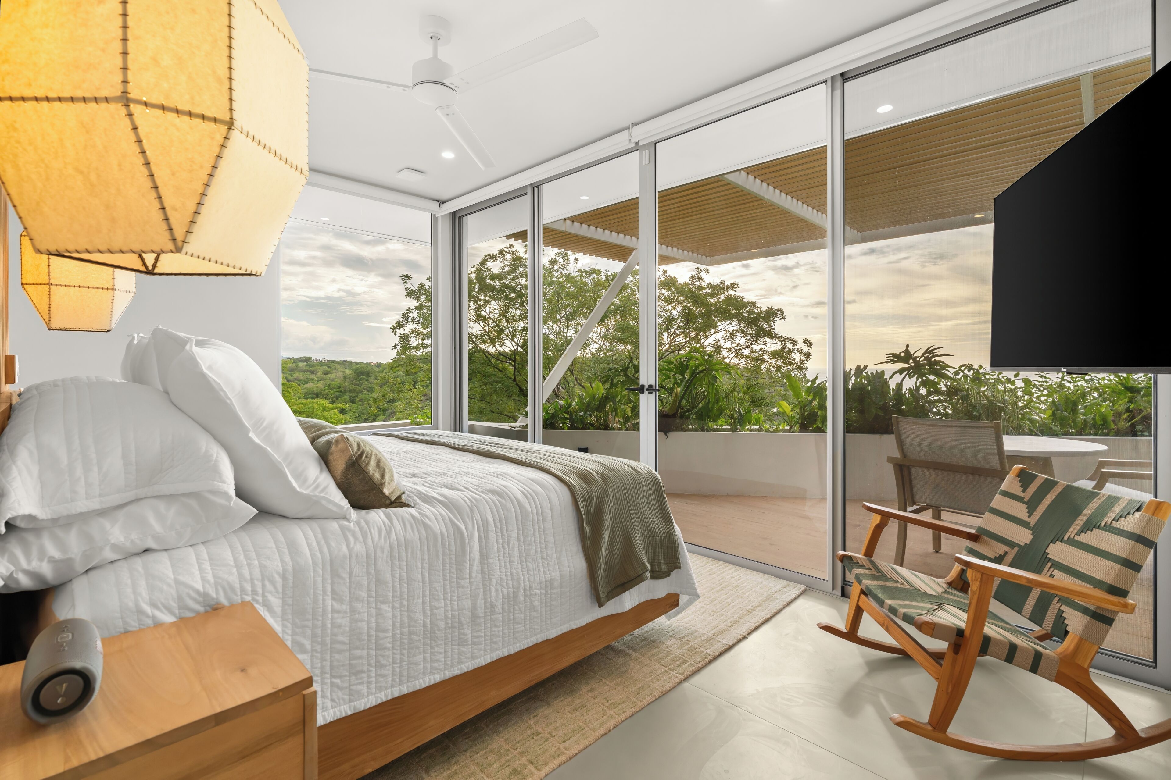 A modern bedroom with a large bed, a wooden nightstand, a chair, and a view of the outdoors through large glass doors.