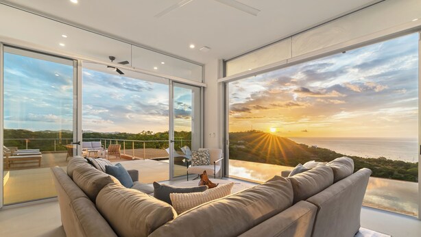 A modern living room with a large sofa, a coffee table, and a view of the sunset over the sea.