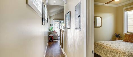 A hallway with wooden flooring, framed pictures on the walls, a ceiling fan, and a bed with a patterned bedspread.