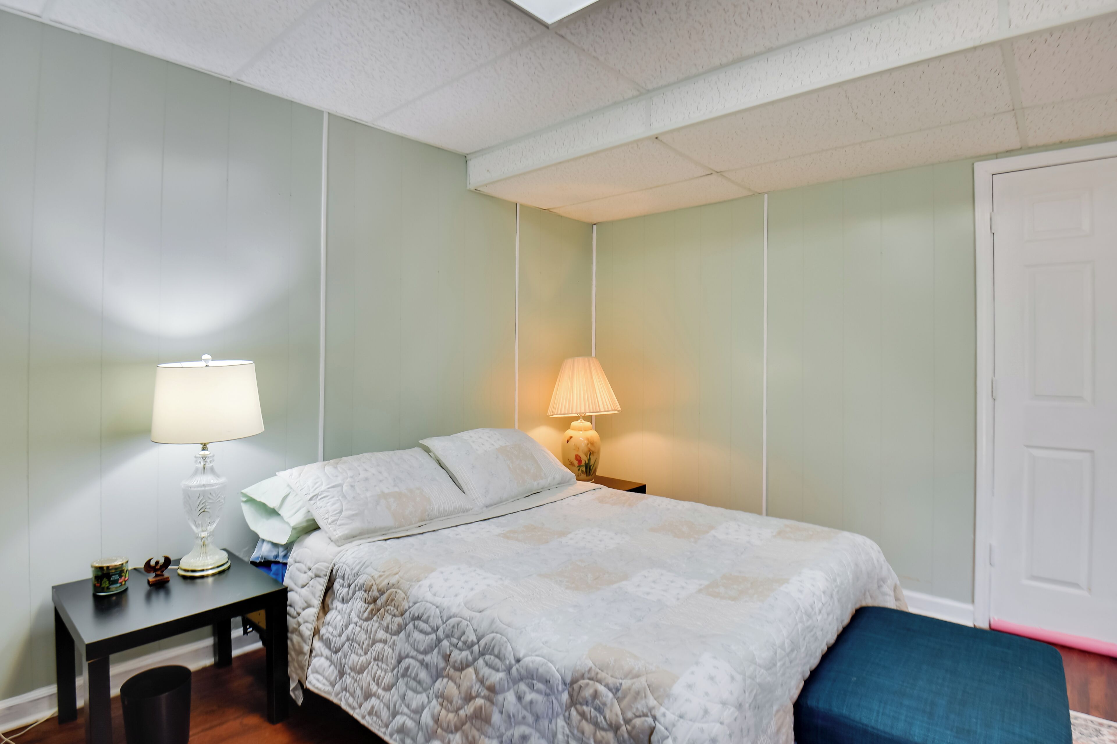 Un dormitorio con una cama, una mesita de noche con una lámpara, una pequeña mesa con una taza y un objeto decorativo, y una puerta.