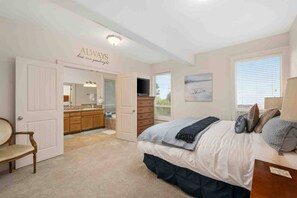 Une chambre à coucher comprenant un lit, une commode, une coiffeuse avec un miroir, une porte donnant sur une salle de bain et une fenêtre avec des stores.