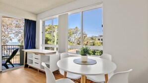 Una habitación luminosa con un escritorio blanco, sillas y un ventanal que ofrece vistas a árboles y un edificio.