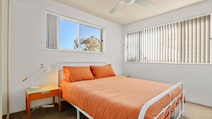 Un dormitorio con cama, mesita de noche, lámpara y dos ventanas.