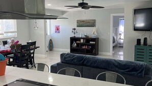 A modern kitchen with a central island, dining area with chairs, a sofa, and a television mounted on the wall.