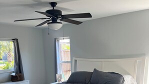 A bedroom with a bed, a ceiling fan, a window with curtains, and a bedside table with a lamp.