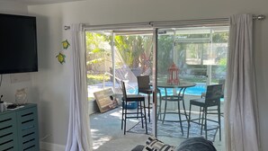 A living room with a sofa, a television, and a sliding glass door leading to an outdoor area with a pool and chairs.