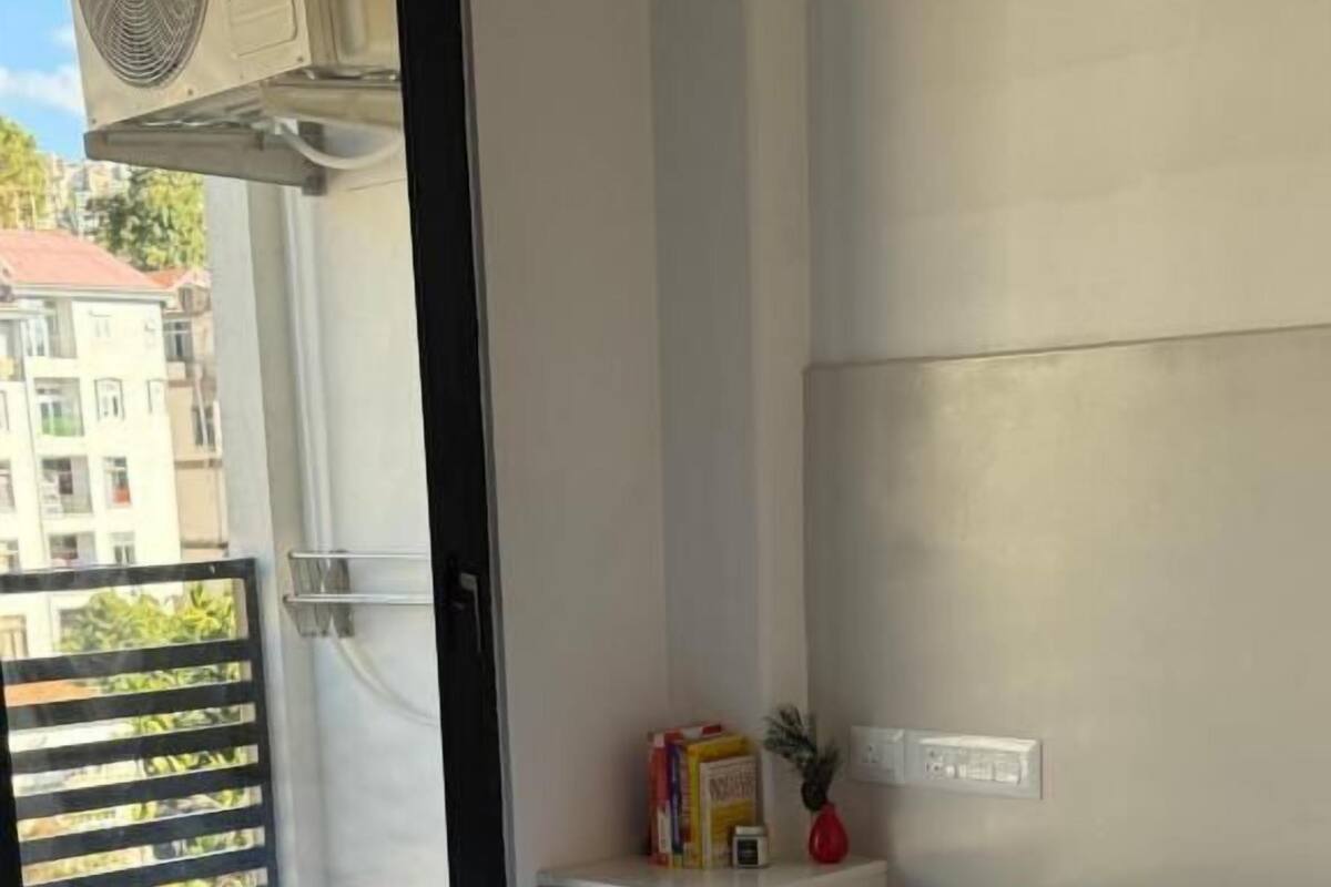 Habitación de hotel con cama, una mesita de noche con lámpara, ventana con vista a edificios y balcón con barandilla.