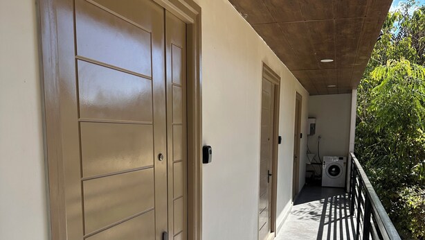 A corridor with beige doors, a balcony with a black railing, and a washing machine visible in the background. - Cornerstone Suites (Aizawl)