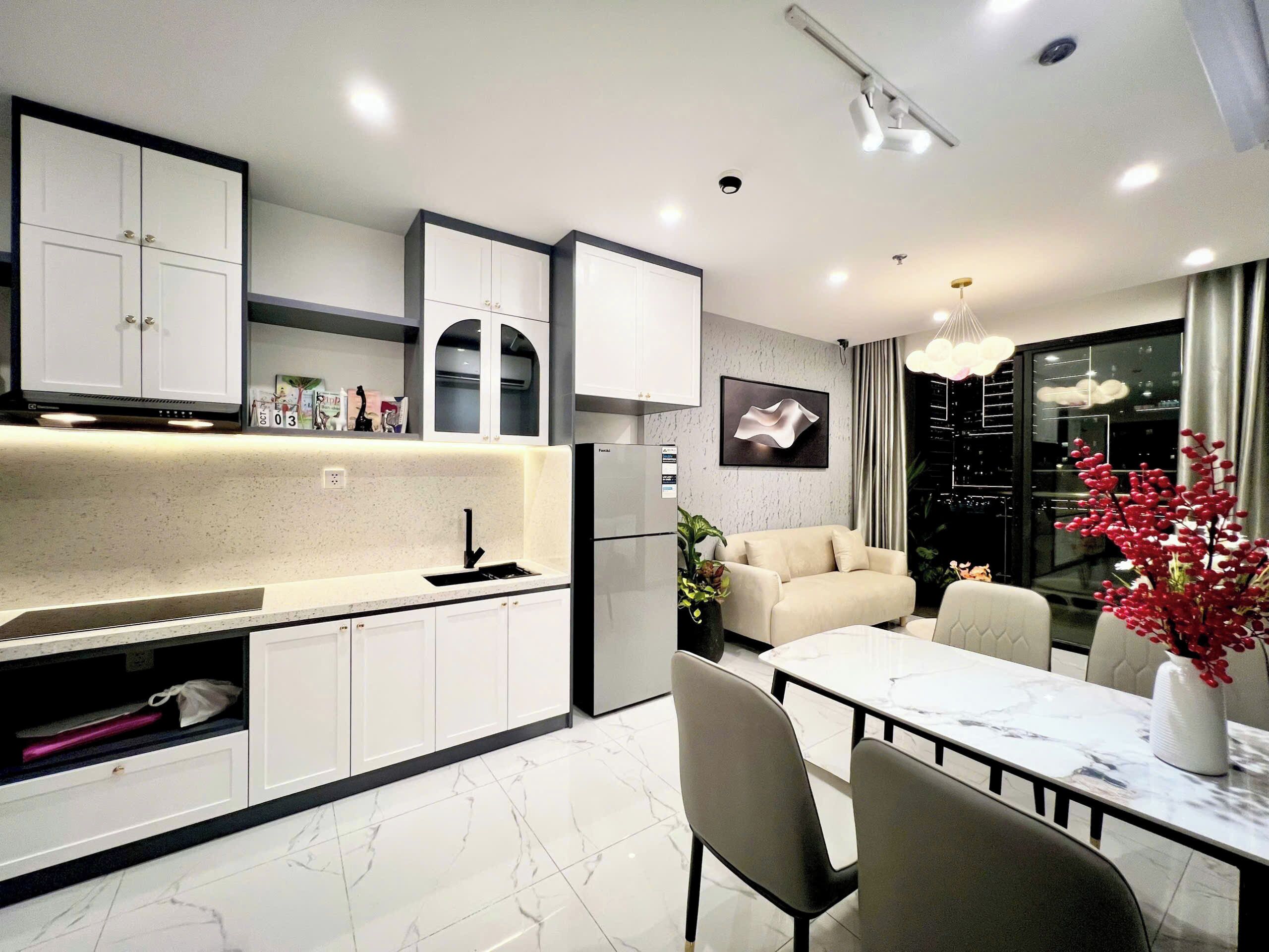 A modern kitchen with white cabinets, a black faucet, and a marble countertop. There is a dining area with a white table and chairs, and a sofa in the background.