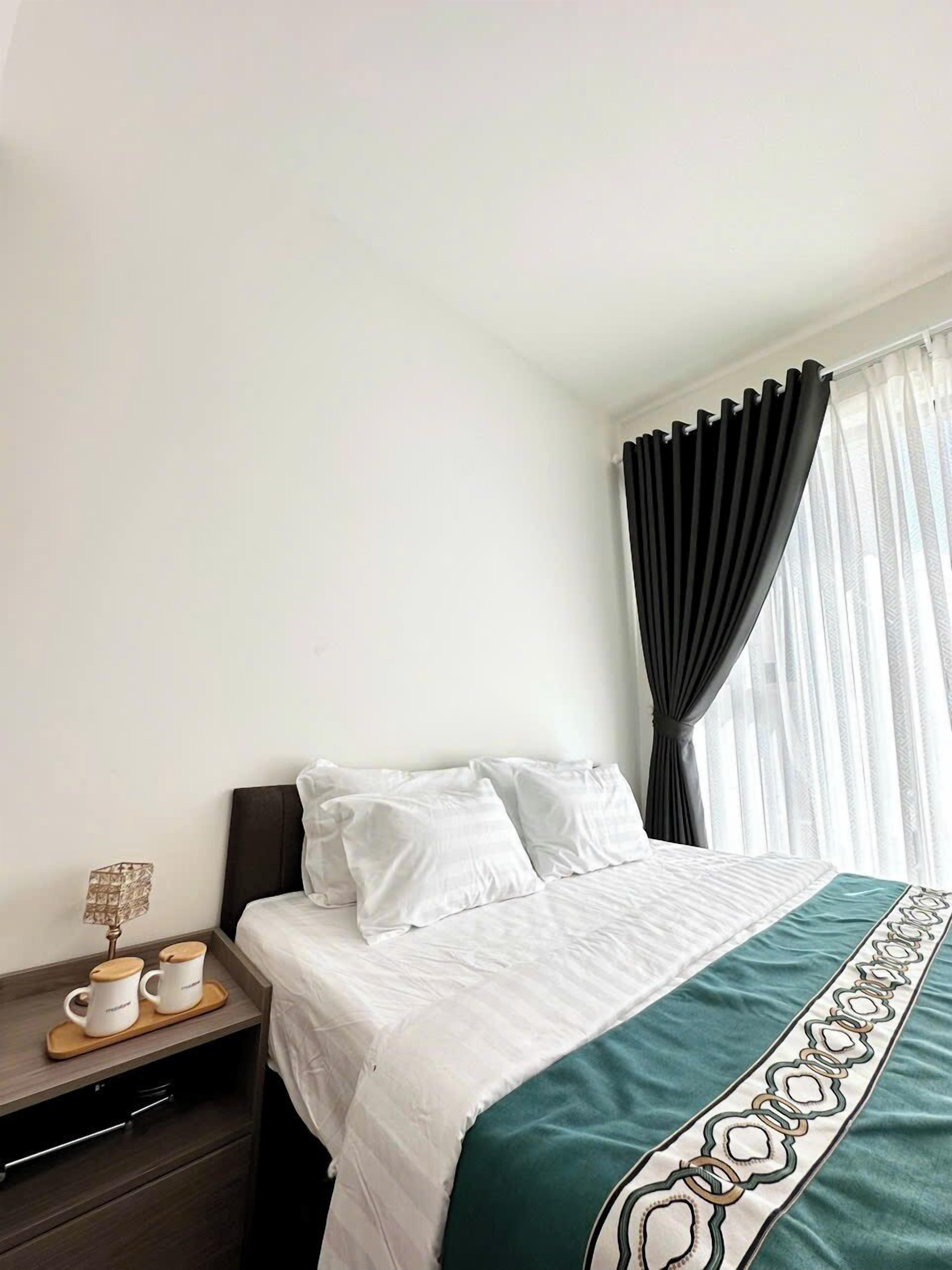 A hotel room with a bed, a nightstand with a lamp and two cups, and a window with curtains.