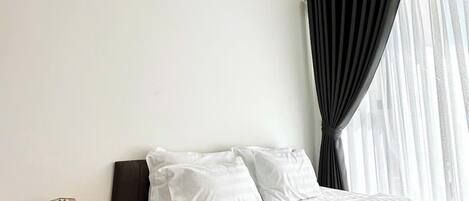 A hotel room with a bed, a nightstand with a lamp and two cups, and a window with curtains.