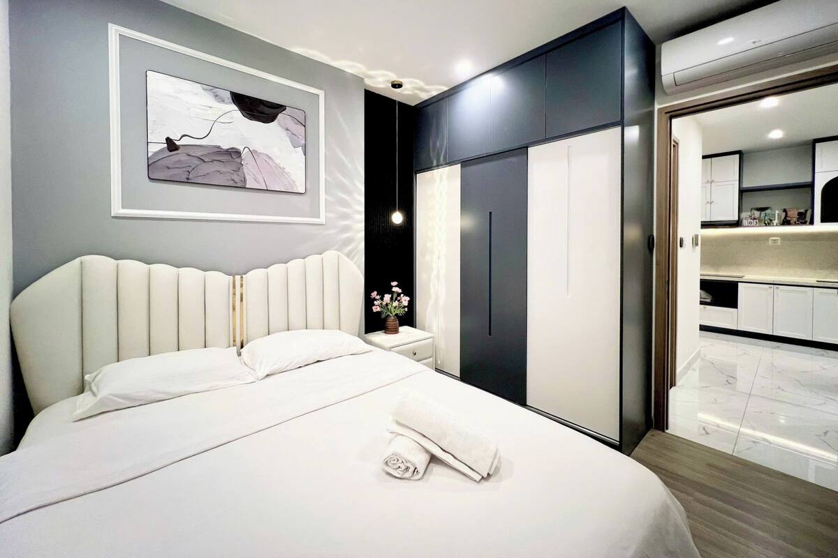 Une chambre moderne avec un grand lit, une table de chevet ornée d’un vase, une armoire et une salle de bain visible à travers une porte ouverte.