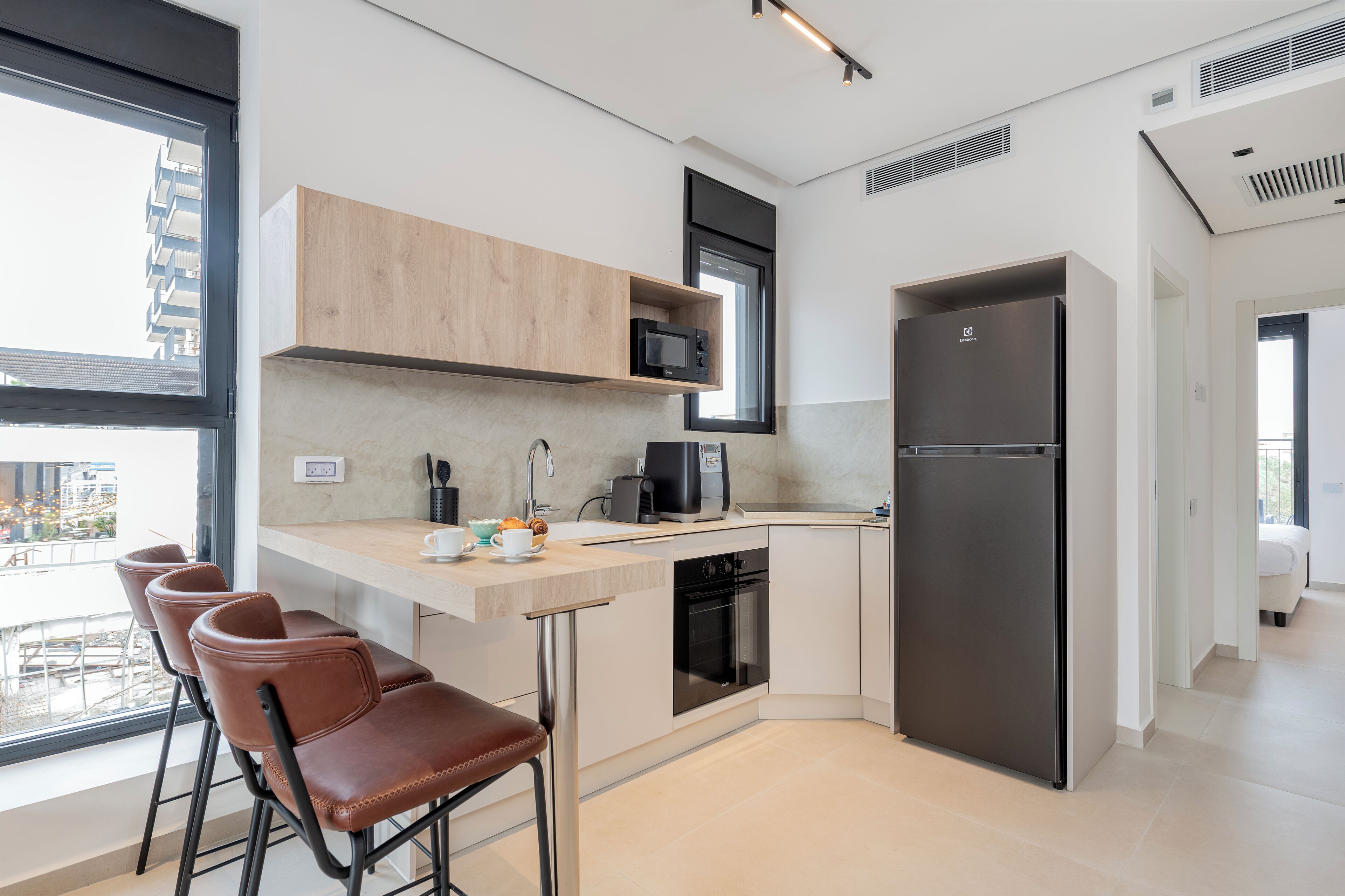 A modern kitchen with a built-in microwave, coffee maker, and refrigerator. There are bar stools and a window overlooking a cityscape.