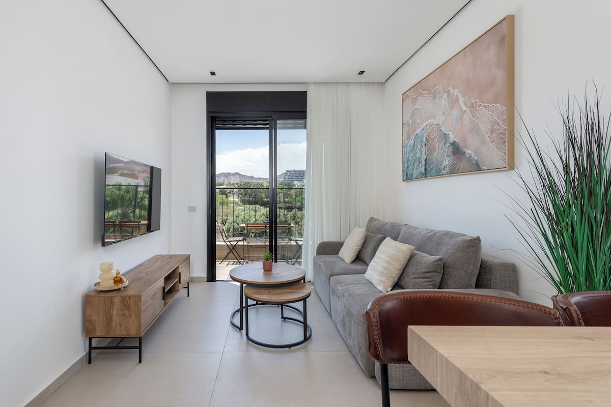 A modern living room with a sofa, a coffee table, a TV, and a balcony with a view.