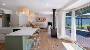 A modern kitchen with a central island, bar stools, a black wood-burning stove, and a large window overlooking a backyard. - Luxurious & spacious retreat near Swan Lake (Sussex Inlet)