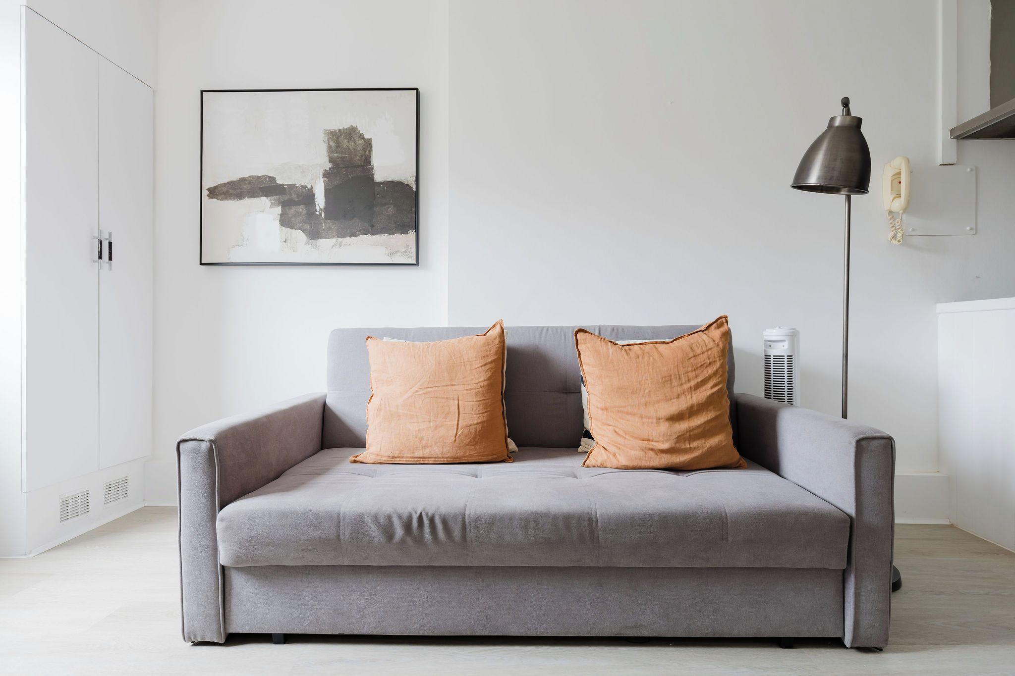 Ein graues Sofa mit zwei orangefarbenen Kissen, eine schwarze Stehlampe und ein gerahmtes abstraktes Gemälde an der Wand.