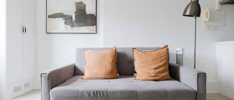 A grey sofa with two orange cushions, a black floor lamp, and a framed abstract painting on the wall.