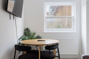 Et værelse med et rundt bord, to stole, et fjernsyn monteret på væggen og et vindue med udsigt til en murstensbygning.