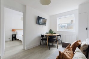 A modern living room with a dining area, a TV mounted on the wall, and a window with a view. - Snug 1BDR City Retreat in West London (Greater London)
