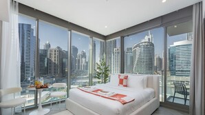 Une chambre moderne avec un grand lit, une petite table où est servi le petit-déjeuner, et une vue sur le paysage urbain grâce à de grandes fenêtres du sol au plafond.