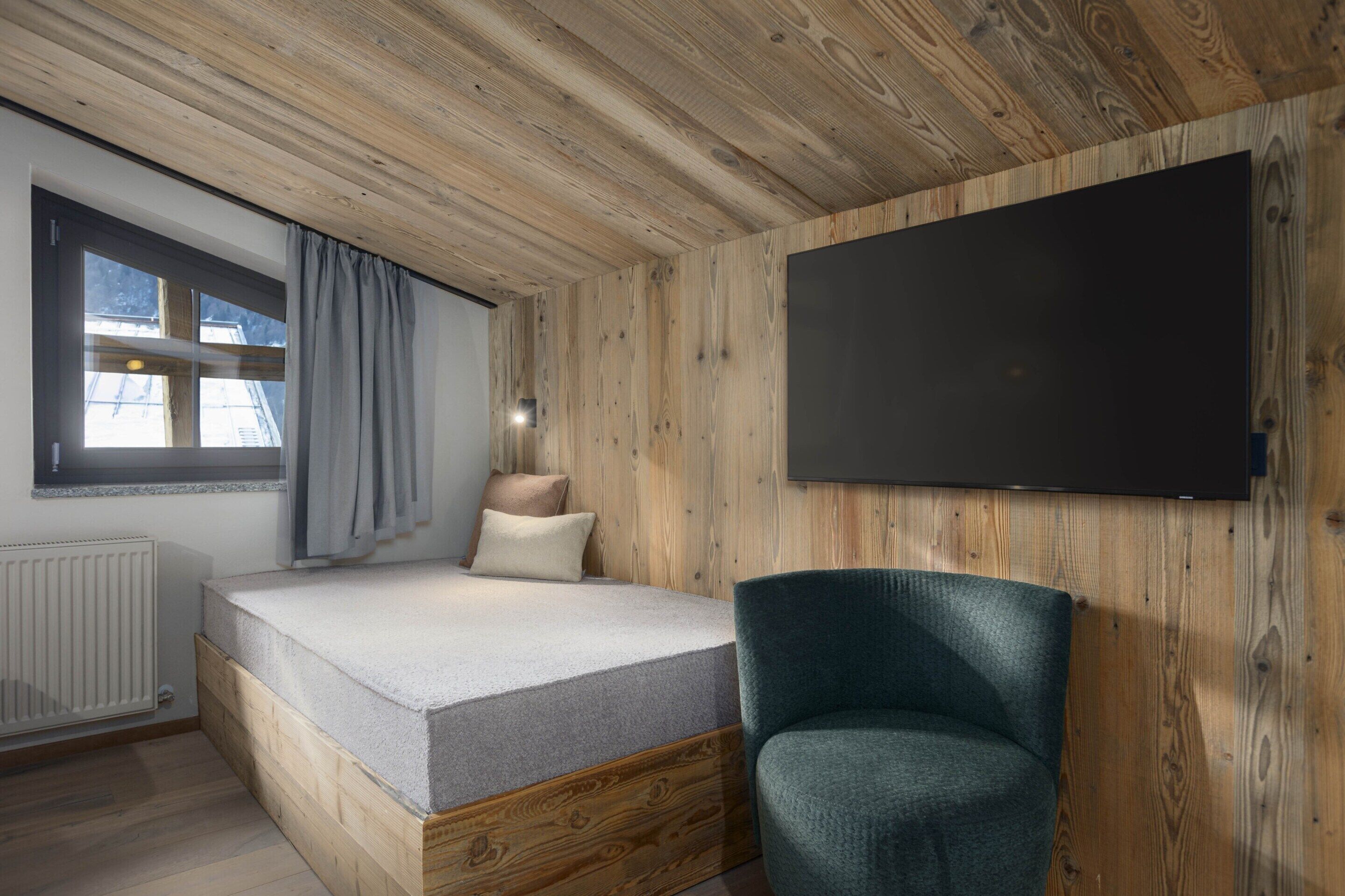Un dormitorio con techo de madera, una cama con colchón y almohadas, una silla, una ventana con cortinas y un televisor de pantalla plana montado en la pared.