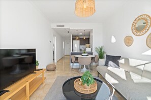 A modern living room with a flat-screen TV on a wooden stand, a round coffee table with a plant, a sofa with cushions, and a dining area in the background. - Modern 4-room apartment with panoramic sea & mountain views (AN)