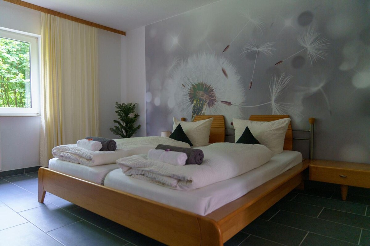 A double bed with neatly folded towels, a bedside table, a potted plant, and a wall with a dandelion pattern.