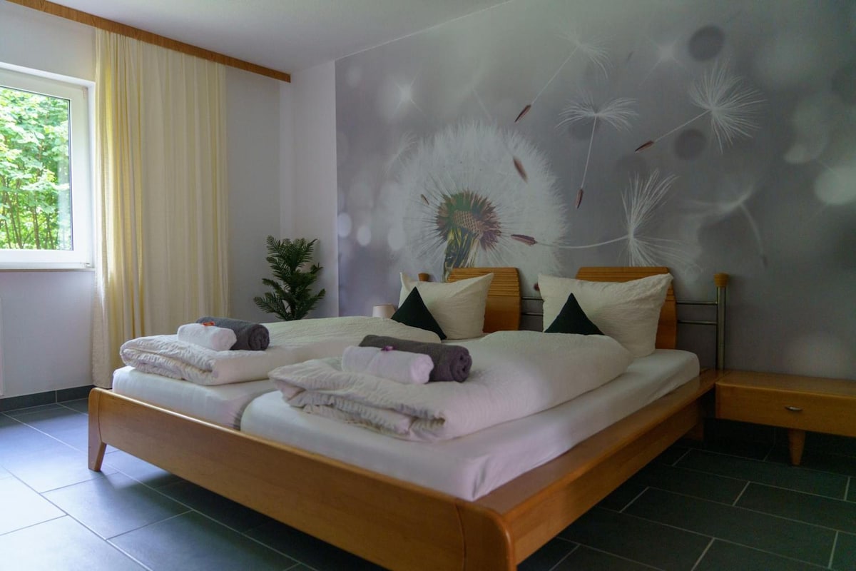 A double bed with neatly folded towels, a bedside table, a potted plant, and a wall with a dandelion pattern.