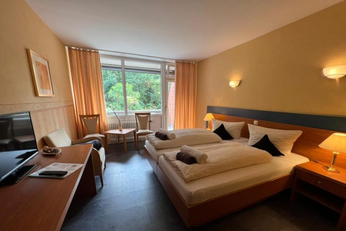 A hotel room with two beds, a desk, a chair, a TV, and a window with curtains.