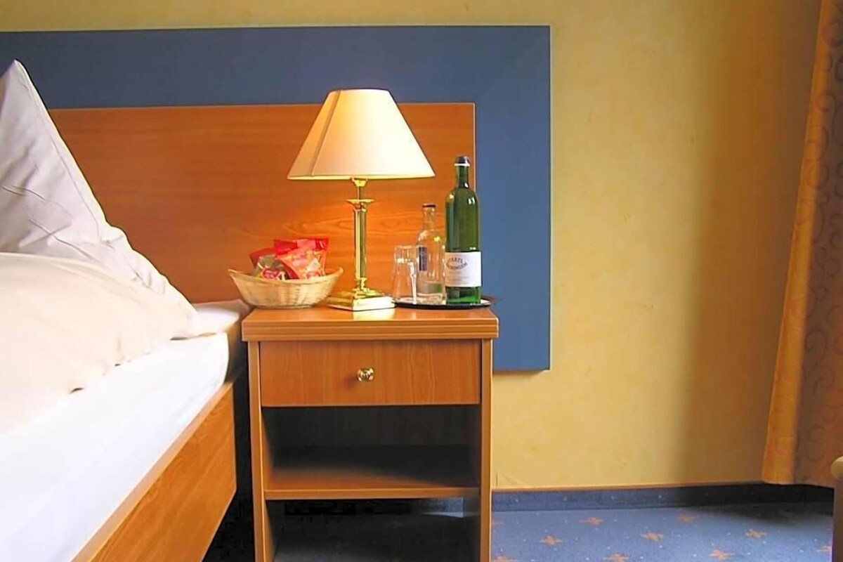 A hotel room with a bed, a nightstand with a lamp, a bottle, and a basket of items.