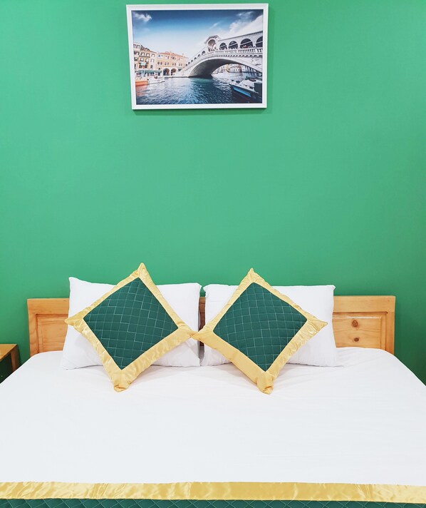 Uma cama com duas almofadas decorativas, cabeceira de madeira e um quadro com uma ponte em uma parede verde.