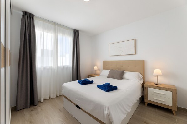 A neatly made bed with two blue towels, a bedside table with a lamp, and a framed picture on the wall. - Merlin 14 V1 Playa Honda (Playa Honda)