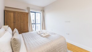 A bedroom with a large bed, wooden wardrobe, and a window with curtains. - Modern 1BDR Near Heathrow with Parking Amenities (Surrey)