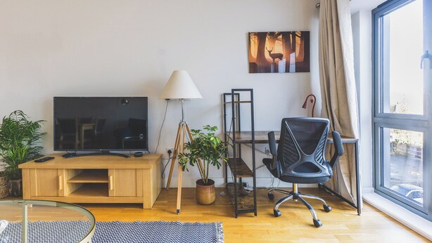 A modern living room with a TV, desk, chair, lamp, and large window. - Modern 1BDR Near Heathrow with Parking Amenities (Surrey)