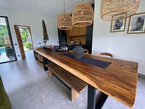 A modern dining area with a wooden table, benches, wicker pendant lights, and a view of the outdoors. - Villa 2ch-Ubud-piscine-Jungle rivière & rizière (UBUD)