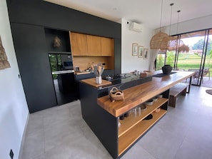 A modern kitchen with a black and wooden island, stainless steel appliances, and a view of the outdoors. - Villa 2ch-Ubud-piscine-Jungle rivière & rizière (UBUD)