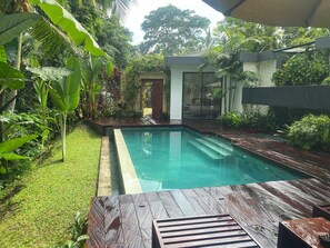 Une piscine dotée d’une terrasse en bois avec des sièges, entourée d’une végétation luxuriante et d’un mur de pierre.
