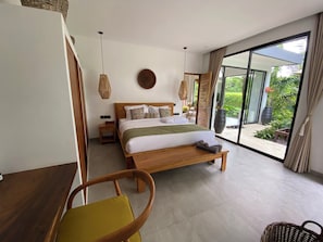 A modern bedroom with a wooden bed, a nightstand, a chair, and a large window offering a view of the outdoors. - Villa 2ch-Ubud-piscine-Jungle rivière & rizière (UBUD)