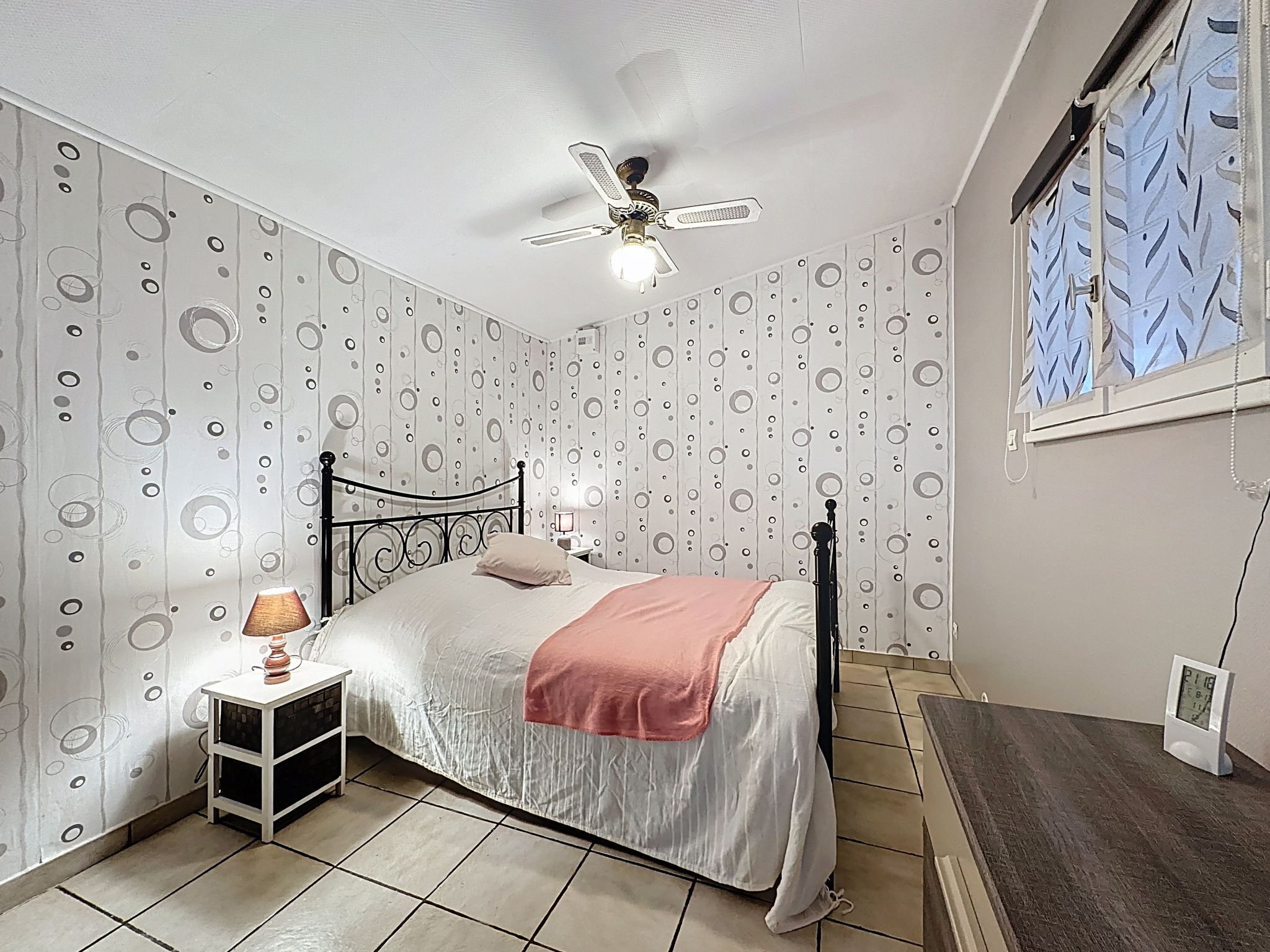 Ein Schlafzimmer mit einem Bett, einem Nachttisch, einem Deckenventilator und einem Fenster mit gemusterten Vorhängen.