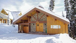 Hirsimökki, jonka perustuksena on kivimuuri, ulkoseinät ovat puuta ja katto on lumen peitossa.