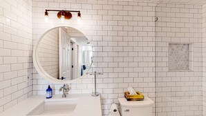 Uma casa de banho com azulejos brancos, um espelho redondo, uma pia, uma sanita e um dispensador de toalhas.