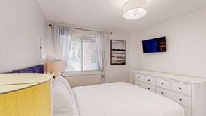 A bedroom with a bed, a bedside table, a lamp, a dresser, a window with curtains, and a wall-mounted TV. - Okanagan - Ice and Cozy #116C (Beaverdell)