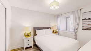 Quarto com cama, duas mesas de cabeceira com luminárias, uma janela com cortinas e um quadro na parede.
