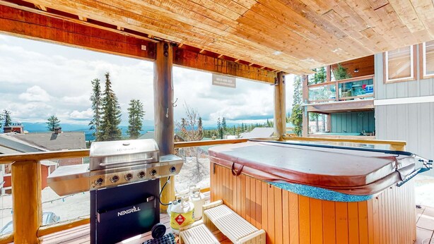 Eine Holzterrasse mit Grill, Whirlpool und Sitzbereich im Freien, mit Blick auf Bäume und Berge.