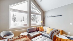 Ein modernes Wohnzimmer mit einer Ledersofa, einem kleinen runden Tisch und einem Sessel. Zwei große Fenster bieten Blick auf verschneite Landschaft.