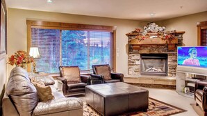 A living room with a stone fireplace, leather sofas, a television, and a large window with blinds. - Okanagan - Snowbanks 4 (Beaverdell)