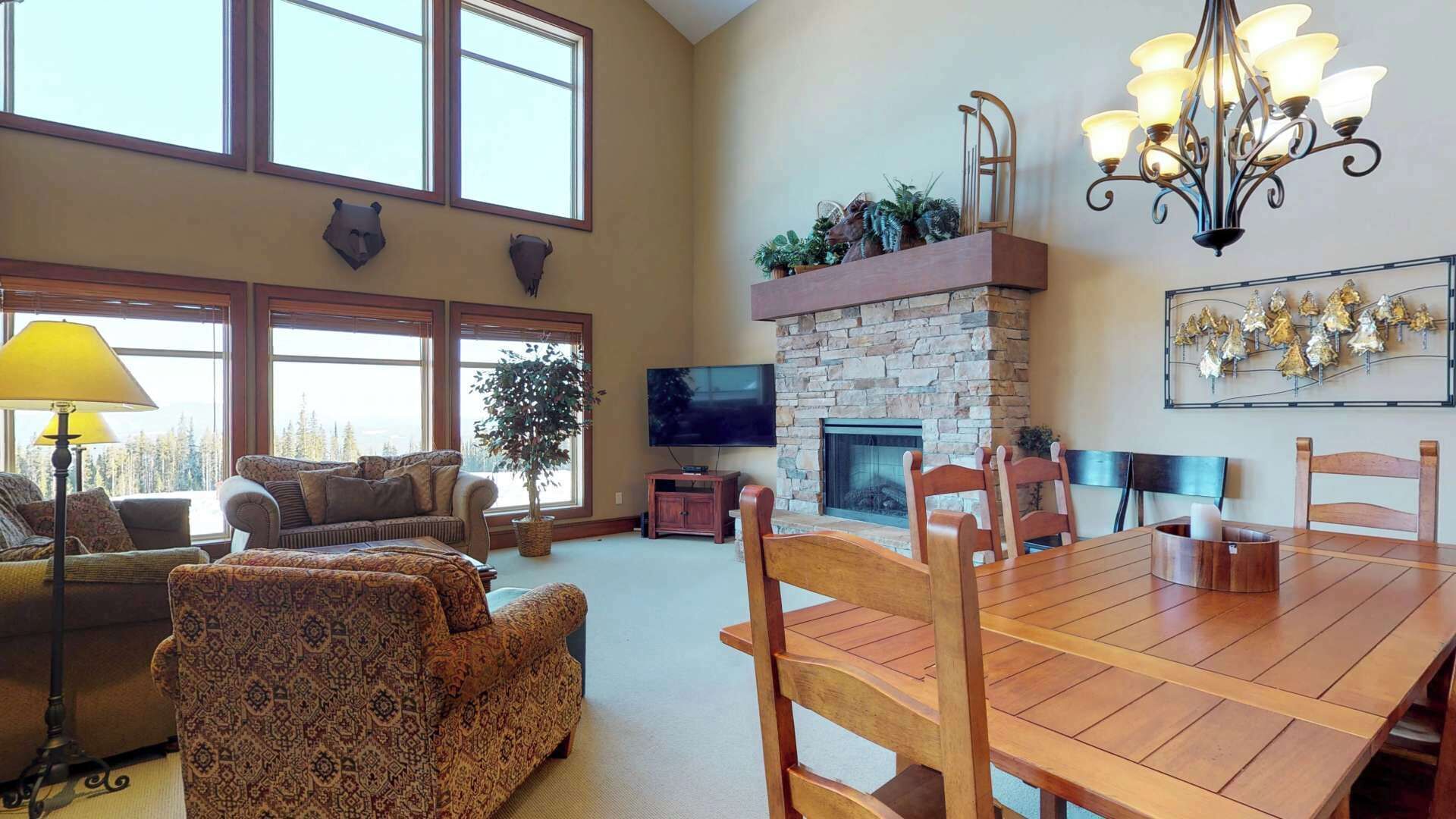 A spacious living room with a stone fireplace, a dining table with chairs, a television, and large windows.