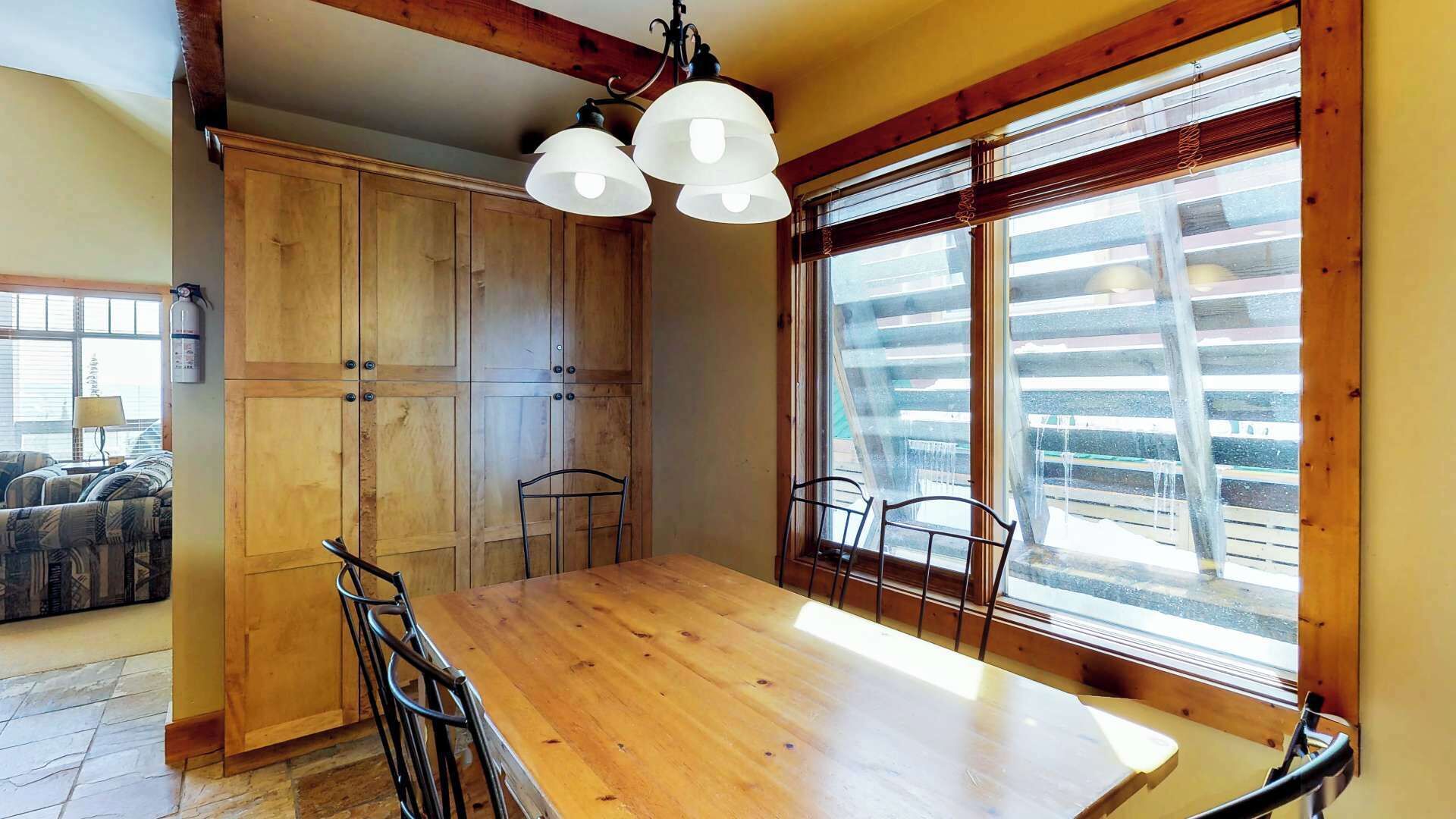 A dining area with a wooden table and chairs, large windows with a view of a balcony, and a ceiling with exposed beams.