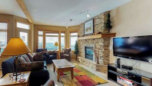 A spacious living room with a stone fireplace, a large flat-screen TV, and a view of the ocean through large windows. - Okanagan - Timber Ridge 17 (Kootenay Boundary E)